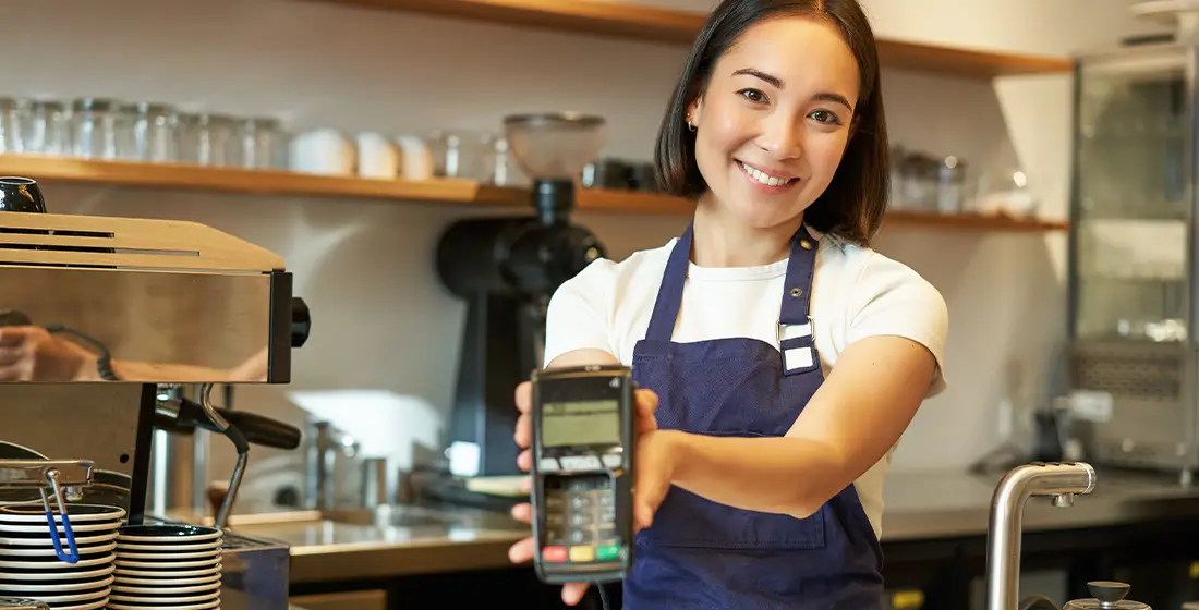Smiling barista in a coffee shop staff holds a credit card machine, ready to process payments with a POS terminal.